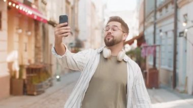 Genç blogcu cep telefonuyla selfie çekiyor, abonelerle çevrimiçi video görüşmesi yapıyor, sosyal medya vlog 'u için hikayeler kaydediyor. Şehir caddesinde yürüyen mutlu adam.