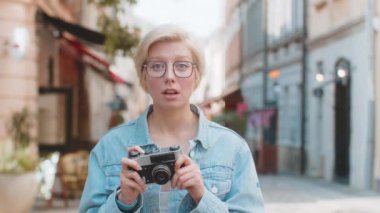 Neşeli beyaz kadın gezginin portresi şehrin caddesinde kamerayla duruyor. Gözlüklü güzel sarışın kadın turist fotoğraf çekerken şaşırdı. Fotoğraf çeken.