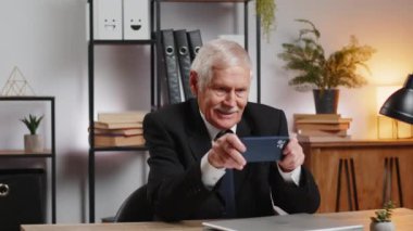 Akıllı telefondan oynayan Kafkasyalı kıdemli iş adamı heyecan verici mobil uygulama ofis masasında oyun kazanma ve atış oyunlarını kutluyor. Profesyonel serbest çalışan adam yeni uygulamanın tadını çıkarsın..