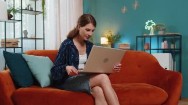 Laptop kullanan Kafkasyalı genç kadın, iyi haberler aldı. Aniden kazanılan zaferle şok oldu. Dairede piyango ikramiyesini kutladı. Neşeli kız evdeki salonda kanepede çığlık atıp dans ediyor.