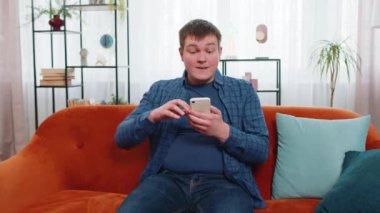 Kafkasyalı genç kazananın elinde akıllı telefon tutarak online bahis oyununun kazanmasına şaşırarak iyi haberler okumasına şaşırdım. Kanepede oturan mutlu erkek. Apartmanda zafer kazanmaktan çok memnun..