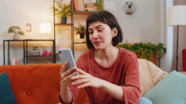Sıradan giysiler içinde gülümseyen genç bir kadın akıllı telefondan evdeki video çağrısında el sallıyor. Kafkasyalı mutlu bir kız cep telefonuyla arkadaşlarıyla konuşuyor. Oturma odasındaki kanepede oturuyor..