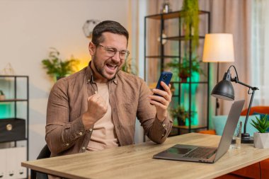 Mutlu genç işadamı akıllı telefon daktilo kullanarak evet diyor, zaferi kutluyor, piyango hedefini kazanıyor olumlu haberler, zafer. Evdeki adam masada oturuyor..