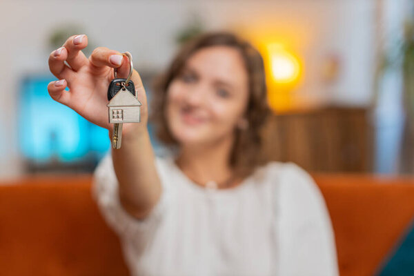 Happy young woman real estate agent lifting hand showing the keys of new home house apartment, buying or renting property, mortgage loan at home indoors. Girl sitting on orange sofa couch. Lifestyles.