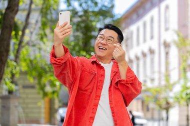 Asyalı orta yaşlı blogcu akıllı telefondan selfie çekiyor, internetten sosyal medya vlog 'u için haber kaydeden abonelerle görüntülü görüşme yapıyor. Şehir caddesinde olgun bir Çinli..