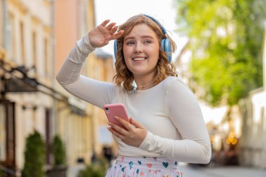 Kablosuz kulaklıklar takmış, dışarıda dans eden akıllı telefon uygulamalarında enerjik müzik dinleyen mutlu, aşırı neşeli kızıl saçlı genç kadın. Şehir caddesinde yürüyen kızıl saçlı kız. Kasaba yaşam tarzları
