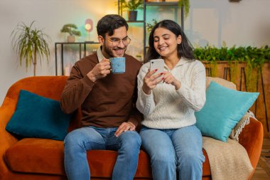 Neşeli genç Hintli çift, akıllı telefondan sosyal medya uygulamalarına göz atarken eğlenceli bir deneyim yaşıyor. Mutlu kız arkadaş ve erkek arkadaş evdeki oturma odasında kanepede konuşuyor.