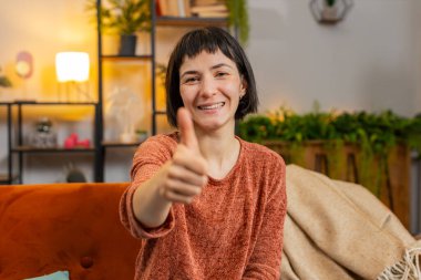Heyecanlı beyaz kadın portresi onaylayıcı bir şekilde kameraya bakıyor, olumlu, iyi haber, olumlu geri bildirim gibi. Evdeki oturma odasındaki kanepede oturan genç kız..
