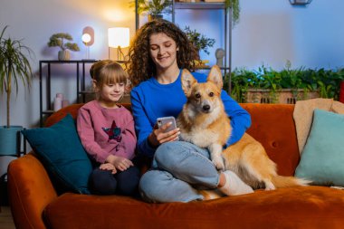 Kanepede akıllı telefon kullanarak Corgi köpeğiyle selfie çeken neşeli anne ve kız çocuğu. Mutlu aile paylaşımı sosyal medya çevrimiçi video görüşmesi hafta sonu boyunca evdeki oturma odasında koltukta oturur.