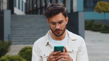 Gülümseyen genç Hintli adam akıllı telefon kullanıyor mesajlar yazıyor internette geziniyor, işi bitiriyor, kameraya bakıyor e-posta okuyor, sosyal medya uygulaması okuyor. Şehir merkezindeki açık havada yaşayan Arap Hintli adam.