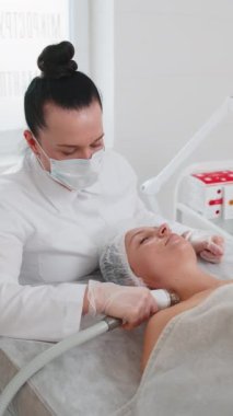 Güzellik uzmanı, kadına yüz ve boyun kaldırma prosedürü yaptırıyor. Kolajen maskesi. Kozmetik uzmanı vakum yüz masajı terapisi için elektrikli ekipman kullanıyor. Cilt bakımı, güzellik tedavisi. Dikey çekim