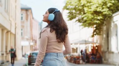 Kablosuz kulaklıklı mutlu Hintli genç kadının arka görüntüsü açık havada akıllı telefondan dans ederek favori disko müziğini dinlerken. Şehir caddesinde yürüyen İspanyol kız. Kasaba yaşam tarzı