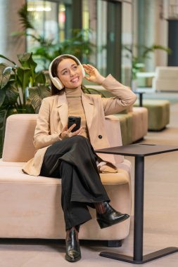 Mutlu, rahat, beyaz, genç iş kadını kulaklık takıyor ve akıllı telefondan favori disko müziğini dinliyor. Formaliteler içinde kadın girişimci dinleniyor, ofis lobisinde oturuyor..