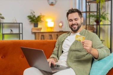 Kredi kartı ve dizüstü bilgisayar kullanan mutlu genç bir adam para transfer ederken, ev içinde alışveriş nakit yemek siparişi veriyor. Kanepede oturan esmer, yetişkin adam..