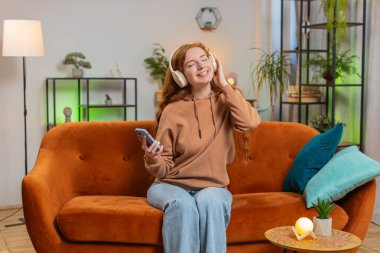 Aşırı neşeli, kablosuz kulaklıklı, evinde koltukta oturup en sevilen disko dansını dinleyen mutlu kızıl saçlı kadın. İnsanlar hafta sonu boş zaman aktiviteleri.