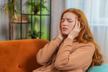 Hasta kızıl saçlı kadın baş ağrısı problemini tedavi etmek için tapınakları ovuyor, tansiyon, migren, stres, ekşitme, evde yüksek tansiyon. Genç yetişkin bir kız odada kanepede..