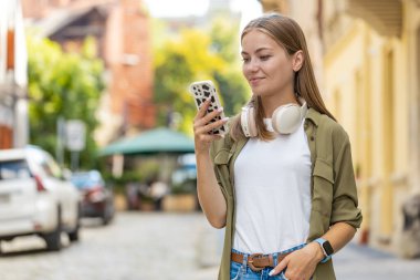 Akıllı telefon kullanarak mesajlar yazan güzel bir genç kadın. İnternetteki sosyal medya uygulamalarına bakarak dışarıda sohbet ediyor. Şehir caddesinde beyaz bir genç kız. Yaşam Tarzları