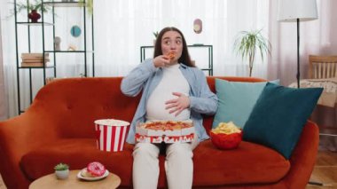 Sağlıksız abur cubur pizza yiyen hamile bir kadın, patlamış mısır gevreği kanepede oturmuş evde televizyon seyrediyor. Hamilelik süresince dairede yağlı yemekler. Açlık, tat tercihlerindeki değişim.