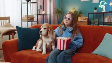 Üç boyutlu gözlüklü, patlamış mısır yiyen ve ilginç TV dizileri, spor oyunları, filmler, çevrimiçi film içerikleri izleyen heyecanlı genç kız. Çocuk, evdeki oturma odasında kanepede av köpeğinin yanında oturuyor..