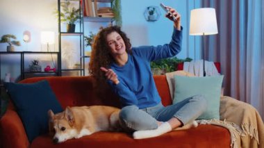 Kıvırcık saçlı neşeli kadın şirin Corgi sosislisini öpüyor ve akıllı telefonuyla evde oturma odasındaki kanepede selfie çekiyor. Mutlu bayan evcil hayvan sever haftasonu köpeklerle vakit geçiriyor.