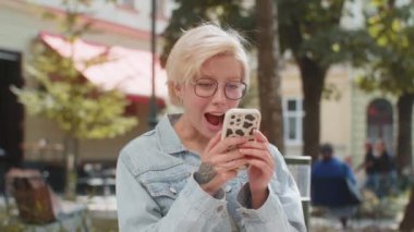 Kafkasyalı genç bir kadın cep telefonuyla kutlama yapıyor iyi haber kazanıyor, piyango büyük ikramiyesi kazanıyor, açık havada dağıtıyor. Şehir caddesinde duran mutlu sarışın bayan turist. Kasaba yaşam tarzları.