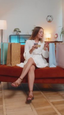 Alışveriş meraklısı genç bir kadın alışveriş poşetleriyle evde oturuyor online sipariş veriyor, kredi kartı ile ödeme yapıyor. İnternet konseptinde indirim. Kanepede beyaz elbiseli bir kız. Dikey.