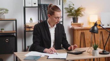 Çalışma saatleri ve finansal grafiklerle uğraşan odaklanmış genç bir ofis çalışanı ofis masasında otururken hesap makinesi kullanıyor. Mutlu stil sahibi işadamı grafikleri karşılaştır ve analiz et.