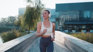 Aktif sporcu kadın koşu maratonu yapıyor, kablosuz kulaklıkla müzik dinliyor. Şehir parkında kardiyo çalışması. Sağlıklı yaşam tarzından zevk alan gülümseyen genç bir kız. Kadın atlet koşucu.