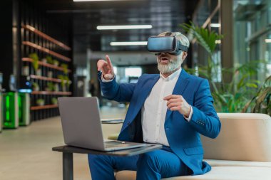 Olgun bir iş adamı yazılım geliştiricisi kulaklık uygulaması kullanarak sunum yapıyor. Sanal gerçeklik 3D 360 videosu izliyoruz. Ofis lobisinde çalışan, VR gözlüklü, beyaz saçlı, takım elbiseli adam..