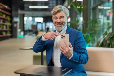 İnternette akıllı telefon uygulaması kullanarak gülümseyen yetişkin işadamı ofis lobisinde çalışan uzaktan kumandayı işaret ediyor. Takım elbiseli adam sosyal medyada e- posta içeriği öğreniyor.