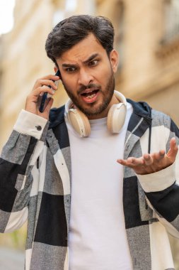 Kızgın kızgın sinirli, sıradan giyinmiş Hintli genç adam akıllı telefonda konuşurken tartışıyor ve dışarıda bağırıyor. Hispanik, şehir caddesinde yürüyen memnuniyetsiz adamı gücendirdi. Kasaba yaşam tarzları.