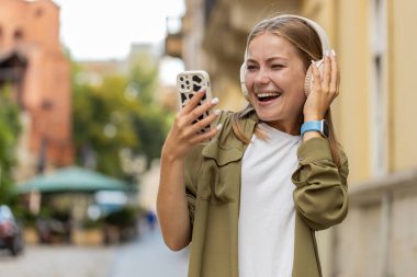 Kablosuz kulaklıklarla gülümseyen, dans eden, favori enerjik disko müziğini dinleyen mutlu genç kadın akıllı telefonları açık havada zaferi kutluyor. Şehir caddesinde duran kız. Kasaba yaşam tarzları.
