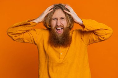 Irritated angry Caucasian man screams from stress tension problems feels horror hopelessness fear surprise shock expresses rage nervous quarrel. Bearded young guy isolated on orange studio background clipart