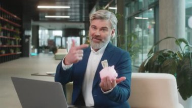 Olgun iş adamı, pembe kumbarayla para biriktirir ve içine dolar atar. Başarı, ekonomi, yatırım, depozito. Modern ofis lobisinde dizüstü bilgisayarlı takım elbiseli adam oturuyor..