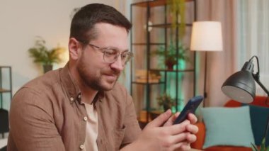 Masada oturan mutlu genç adamın portresi evde gülümseyen cep telefonu kullanıyor. Guy Freelancer akıllı telefon sosyal medya uygulamaları üzerinden online film izlerken mesajlarını paylaşıyor