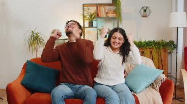 Heyecanlı genç çift dans ediyor, ritme ayak uyduruyor, ellerini ovuşturuyor, dublaj dansı yapıyor. Neşeli mutlu Hintli aile evdeki oturma odasındaki kanepede oturarak eğleniyor..