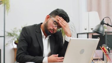 Tired ill Indian businessman suffering from headache problem tension and migraine, stress at home office desk workplace. Bearded freelancer broker worker man works on laptop notebook. Overworking