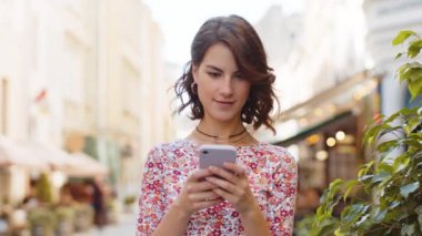 Gülümseyen mutlu genç bir kadın akıllı telefon kullanıyor mesajları cevaplıyor internetteki görüntülü sosyal medya uygulaması ile sohbet ediyor. Kız turist ellerinde telefonla şehir sokaklarında yürüyor.