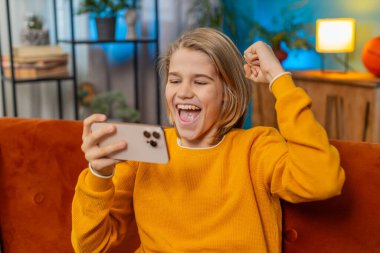 Akıllı telefondan oyun oynayan mutlu beyaz çocuk neşe ve heyecan yaşıyor. Çocuk oyunu kazanır, evdeki kanepede mutlu bir şekilde zıplar, 