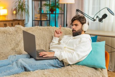 Kablosuz kulaklıklı mutlu Hintli genç adam kanepeye oturmuş dizüstü bilgisayar üzerinde çalışırken disko müziği dinliyor. Hafta sonu evde serbest çalışan sakallı adam eğleniyor..