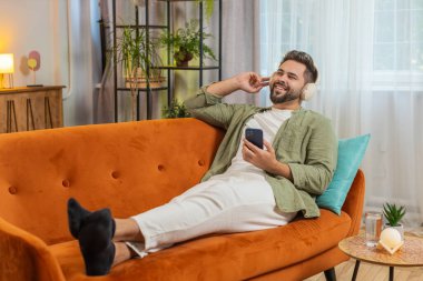 Kablosuz kulaklıklı, akıllı telefonlu mutlu genç adam evinde kanepeye uzanmış, en sevilen enerjik disko dans müziğini dinliyor. Hafta sonu eğlence aktiviteleri.