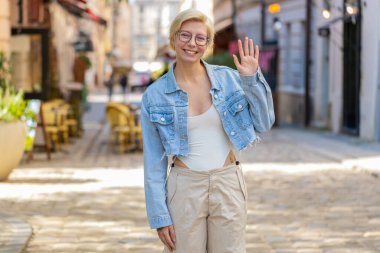 Neşeli genç bayan turist kameraya gülümsüyor, el sallıyor, el sallıyor merhaba, selam, selam, hoşçakal, misafirperver bir yüz ifadesiyle hoş geldiniz. Şehir caddesinde gözlüklü bir kadın.