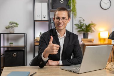 Mutlu, aşırı neşeli, heyecanlı, genç, Kafkasyalı adam. İnternetteki zafer konseptini kutlamak için dizüstü bilgisayarla heyecanlı. İç işlerindeki masada oturan çifte başparmakları gösteren kameraya bakıyor. Takım elbiseli adam.