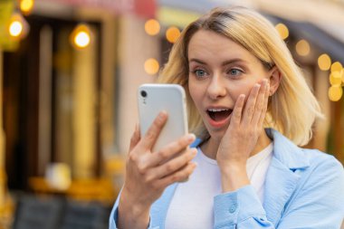 Aman Tanrım. Vay canına. Heyecanlı mutlu, neşeli genç bayan akıllı telefon kullanarak ekranda kutlama yaparak iyi haber kazanıyor. Şehir caddesinde cep telefonu olan beyaz kadın bir gezgin.