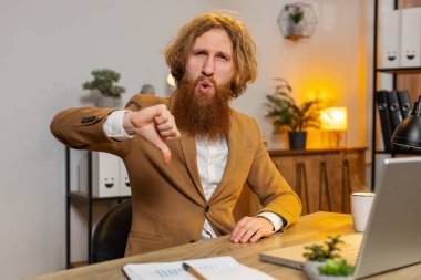 Hoşlanmamak. Kafkasyalı işadamı, ofisteki dizüstü bilgisayarda çalışırken, hoşnutsuzluğunu, tatminsizliğini ve kötü çalışmalarını gösteren işaret işaretini gösteriyor. Hoşnut olmayan kızıl saçlı serbest çalışan.