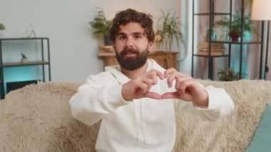 Ben de seni seviyorum. Oturma odasındaki kanepede oturan mutlu Hintli genç adamın portresi sevgiyi simgeler, kameraya kalp işaretini gösterir, romantik hisleri samimi ve olumlu duygular ifade eder. Minnettarlık.