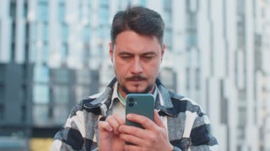 Happy şoke olmuş Kafkasyalı yetişkin turist gündelik giysiler içinde ağzı açık bir şekilde zaferi kutluyor cep telefonlarıyla iyi haberler taşıyor, mesajlar kazanıyor şehir merkezinin dışında başarılar diliyor.
