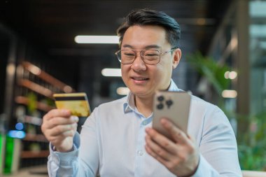 Modern ofis lobisinde online alışveriş ödemeleri için banka kartı ve akıllı telefon kullanan mutlu Çinli işadamı. İş anlaşması. Gülümseyen adam cep telefonu E-bankacılık uygulama servisiyle alışveriş yapıyor..