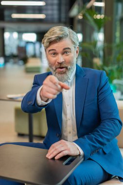 Seni seçiyorum. Kafkasyalı orta yaşlı işadamı kamerayı işaret ederek, mutlu bir ifadeyle bakarak, işe alma seçimini yapıyor. Resmi takım elbiseli mutlu adam modern ofis otel lobisinde oturuyor..