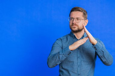 Kafkasyalı orta yaşlı bir adamın mola verme hareketi, sınır veya dur işareti göstermesi, baskı yok, daha fazla zamana ihtiyacım var, dinlenmek, dinlenmek, yardım. Genç yetişkin adam mavi arka planda izole edilmiş